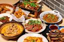 A table filled with various Asian dishes, including sliced meat in rich orange sauce, fried chicken bites, stir-fried vegetables with herbs, a dish of cooked ground meat with peppers, and grilled meat on a platter, all garnished with cilantro.