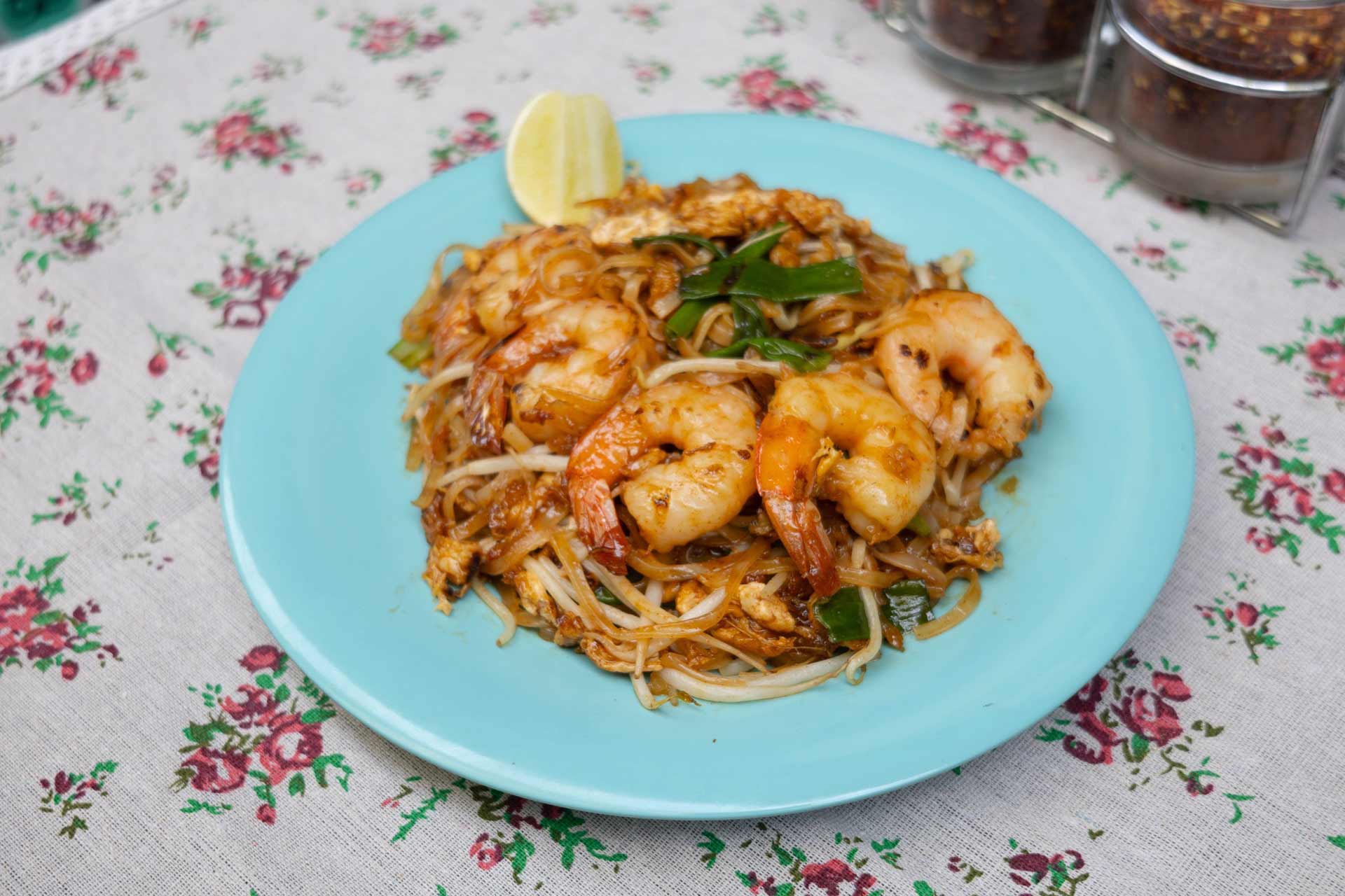 A light blue plate with pad thai topped with shrimp, bean sprouts, green onions, and a wedge of lime, placed on a floral-patterned tablecloth.