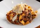 A plate of wide pappardelle pasta topped with a rich meat ragu sauce, wilted greens, and garnished with shaved Parmesan cheese, served on a white dish.
