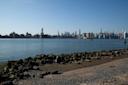 View of a city skyline under a clear blue sky, seen across a body of water from a rocky shoreline. Prominent skyscrapers are silhouetted in the distance, reflecting modern urban architecture.
