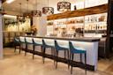 A modern bar interior featuring a row of blue cushioned stools along a sleek counter. The backlit shelves display various bottles. Pendant lights hang above, and a floral arrangement adds a touch of color to the minimalist decor.
