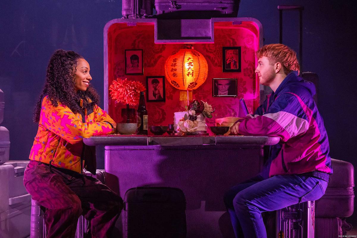 Two people sit across from each other at a small, decorative table with red lighting and a glowing lantern between them, smiling and engaged in conversation. The setting is cozy, with framed photos and flowers on the table.