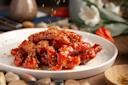 A plate of glazed chicken wings sprinkled with herbs on a white plate. The wings have a shiny, rich red sauce. In the blurred background, there are flowers and smooth stones, adding an earthy ambiance to the setting.