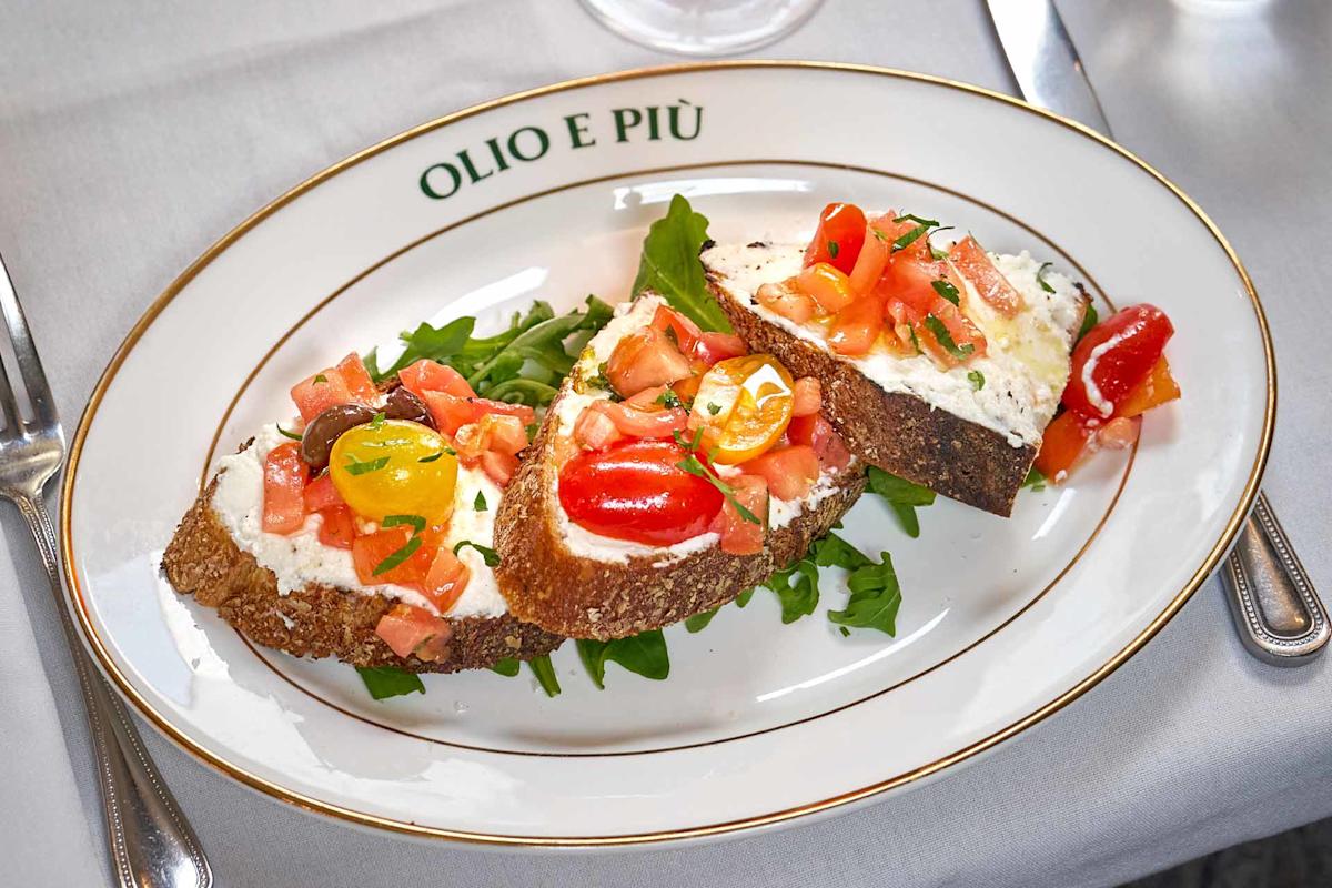Three slices of bread topped with ricotta cheese, chopped tomatoes, and herbs sit on a bed of arugula on a white plate labeled “OLIO E PIÙ.” Silverware and a white napkin are visible beside the plate.