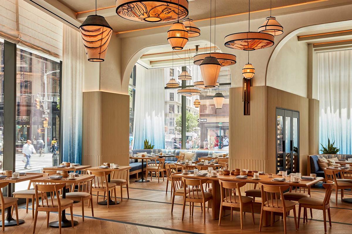 Modern restaurant interior with wooden tables and chairs, large windows letting in natural light, decorative pendant lights, and softly colored walls. Tables are set for dining, and people can be seen outside through the windows.