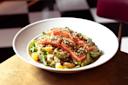 A bowl of salad featuring sliced, seasoned salmon on top of arugula, orange segments, and avocado. The dish is garnished with seeds and herbs, set on a wooden table with a blurred checkered floor in the background.