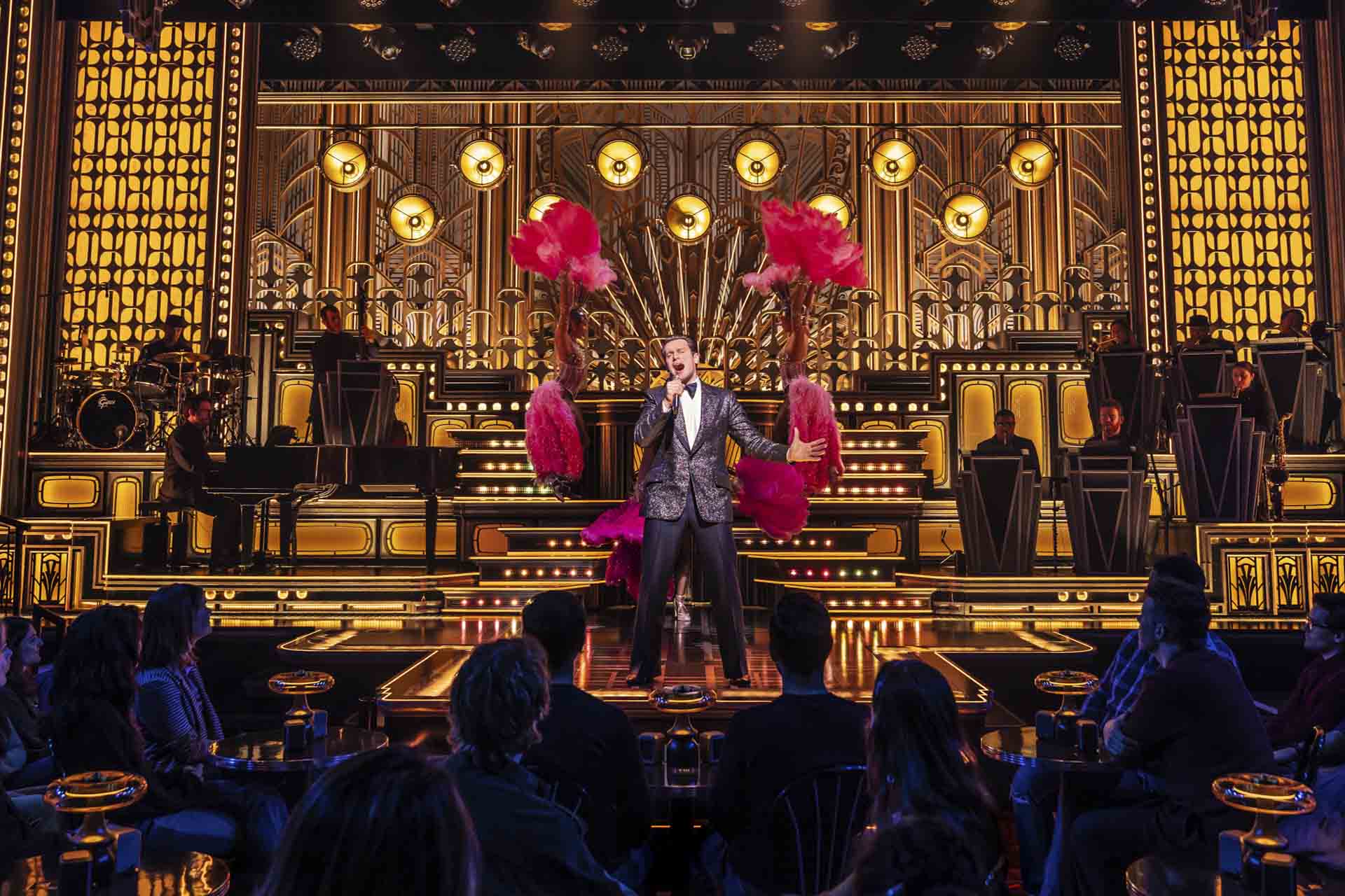 A singer in a sparkling suit performs on stage with showgirls holding pink feather fans behind him, surrounded by Art Deco-style gold and black decor, while an audience watches.