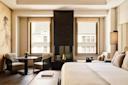 A modern, elegant hotel room with a large bed, sitting area, fireplace, tall windows with city views, and neutral-toned decor. Natural light fills the space, highlighting the cozy and minimalist design.