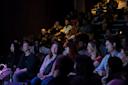 A diverse audience sits in dim lighting, focused on a stage. The front row is illuminated, showing people smiling and engaged. The backdrop is a shadowy theater filled with more attendees.