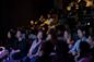 A diverse audience sits in dim lighting, focused on a stage. The front row is illuminated, showing people smiling and engaged. The backdrop is a shadowy theater filled with more attendees.