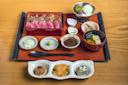 A wooden table displays a Japanese meal set including sliced steak, rice, miso soup, dipping sauces, and small dishes of pickled vegetables and tofu. A dessert with green tea ice cream is in a bowl. A small spoon rests on the tray.