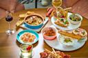 A wooden table set with various appetizers, bread, meatballs, olives, shrimp, and desserts, along with glasses of white and red wine; two people’s hands are partially visible.