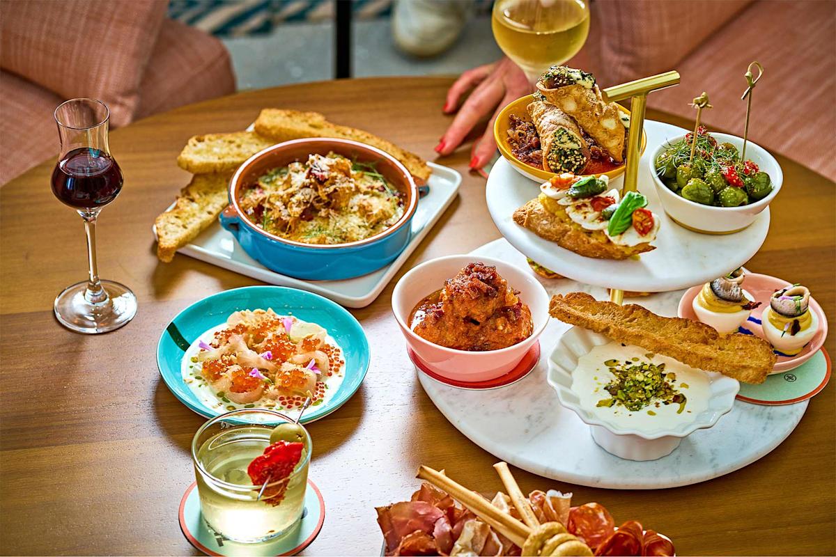 A wooden table set with various appetizers, bread, meatballs, olives, shrimp, and desserts, along with glasses of white and red wine; two people’s hands are partially visible.