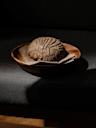 A round loaf of dark bread with a textured crust sits on a cloth napkin in a wooden bowl, placed on a dark surface with dramatic lighting and deep shadows.