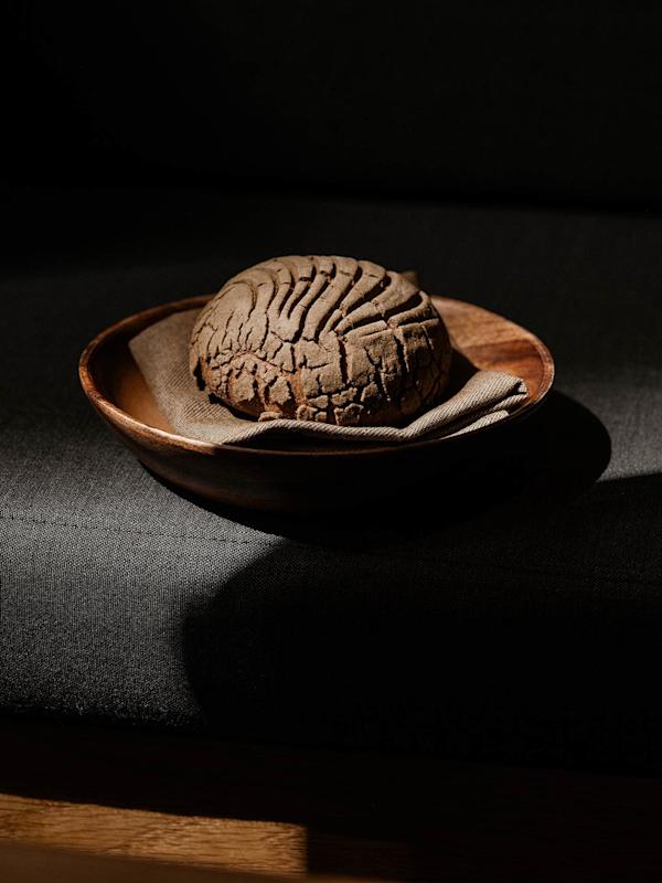 A round loaf of dark bread with a textured crust sits on a cloth napkin in a wooden bowl, placed on a dark surface with dramatic lighting and deep shadows.