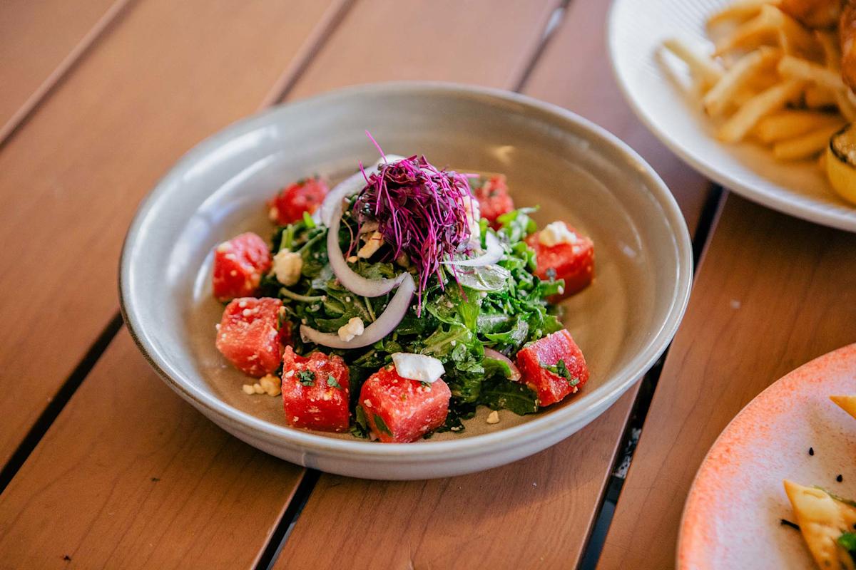 A bowl of salad with watermelon cubes, mixed greens, sliced onions, crumbled cheese, and a garnish of purple sprouts, served on a wooden table.