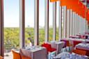 Modern restaurant with large floor-to-ceiling windows overlooking a green park and city skyline. Tables are set with white tablecloths and colorful chairs, and orange ceiling panels add a vibrant touch.