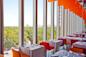 Modern restaurant with large floor-to-ceiling windows overlooking a green park and city skyline. Tables are set with white tablecloths and colorful chairs, and orange ceiling panels add a vibrant touch.