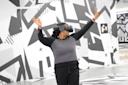 A person wearing a virtual reality headset stands with arms raised in front of a black-and-white geometric mural. The person wears a patterned sweater and black pants, appearing to interact with the VR environment.