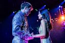 A couple in sparkly outfits gazes into each other's eyes while holding hands on a stage. Bright colorful lights shine in the background, creating a romantic and glamorous atmosphere.
