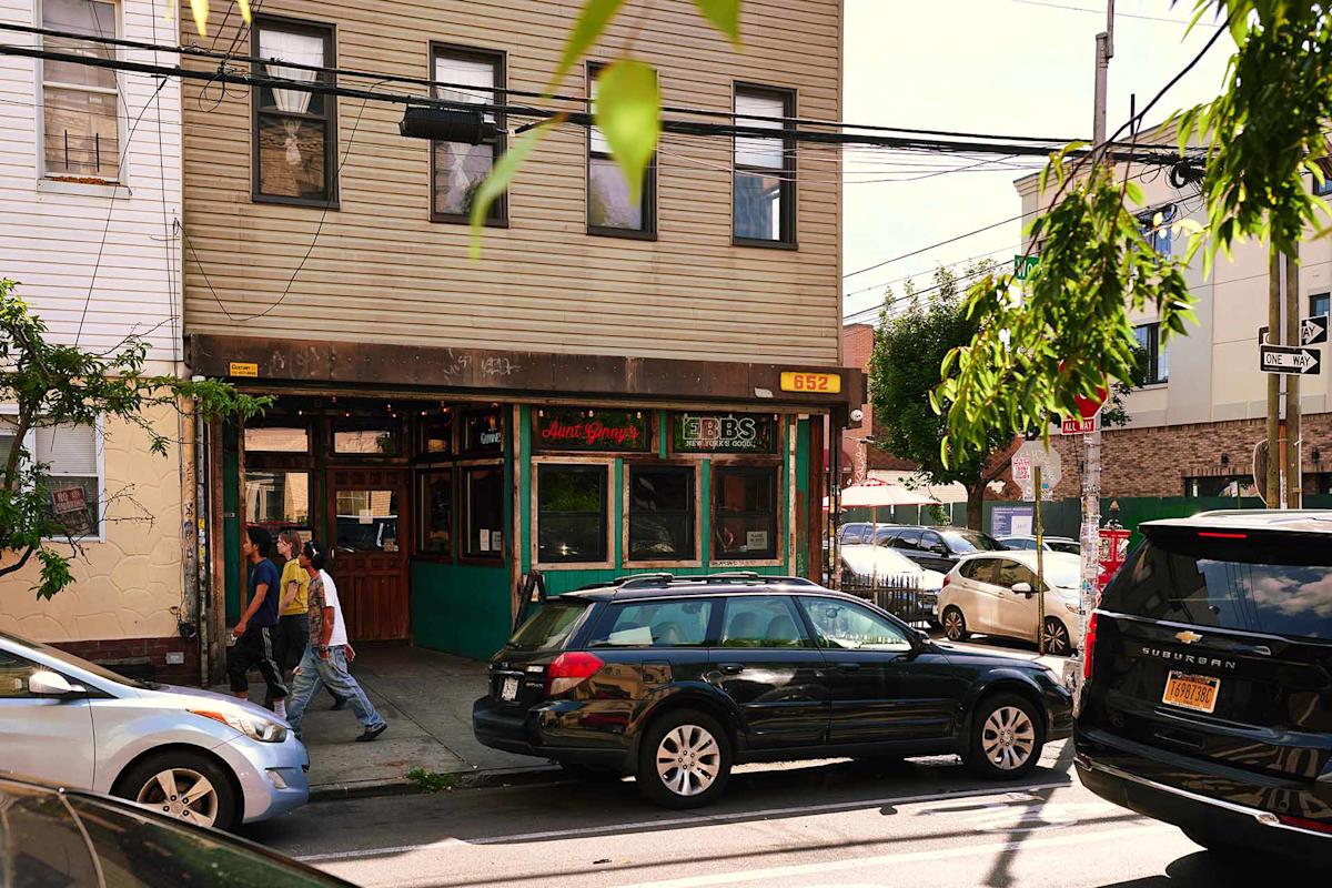 Two people walk past Aunt Ginny’s bar on a sunny street. Parked cars line the curb, and green trees partially frame the view. The building has tan siding with windows above the storefront.