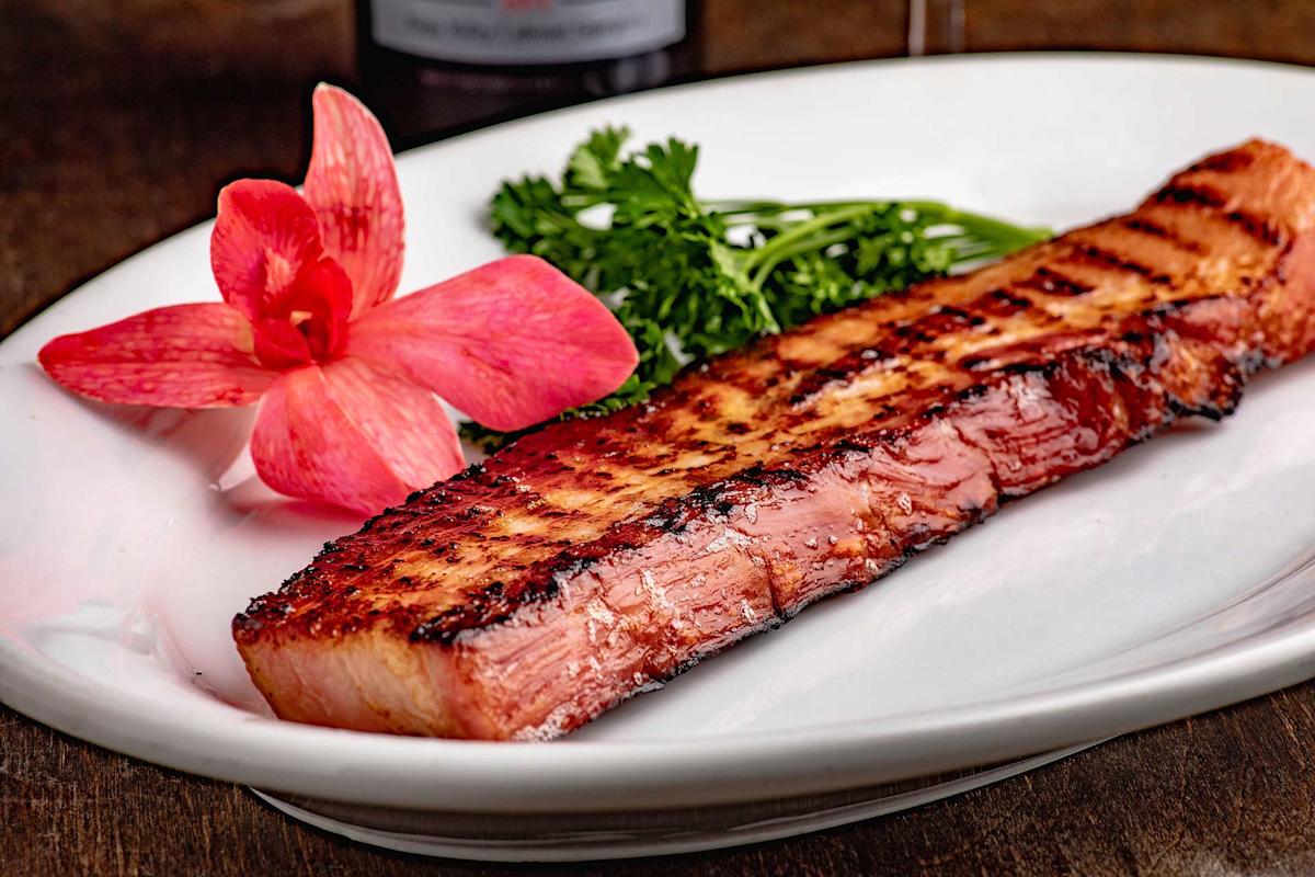 A grilled steak served on a white plate, garnished with a sprig of parsley and a pink flower.