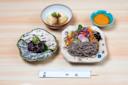 A Japanese meal with soba noodles, colorful vegetables, a dish of cooked eggplant, tofu topped with green onions, and a small bowl of orange seasoning, arranged neatly on a light wooden table with chopsticks.