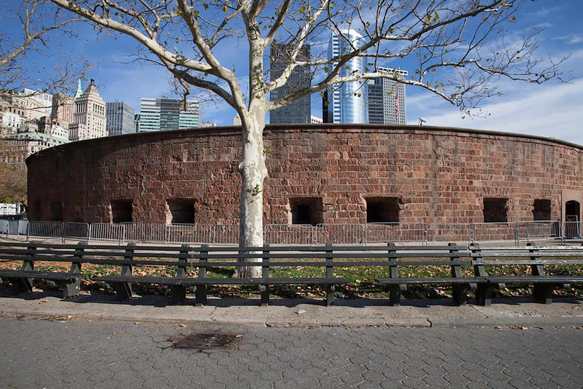 A round, red-brick historic fort with small rectangular windows, surrounded by metal barricades and leafless trees, stands in a city park with modern skyscrapers in the background.