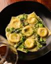 A black plate of round ravioli in a creamy sauce, garnished with green peas, asparagus, microgreens, and cracked pepper, with a blurred glass of wine in the foreground.