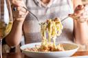 A person twirls creamy fettuccine pasta with a fork and spoon over a white bowl, with a glass of white wine visible on the table.