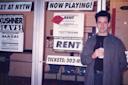 A person holding a drink stands in front of a theater displaying "Rent" posters, with "Now Playing!" and "Sold Out" signs. The background includes glass doors and additional theater advertisements.