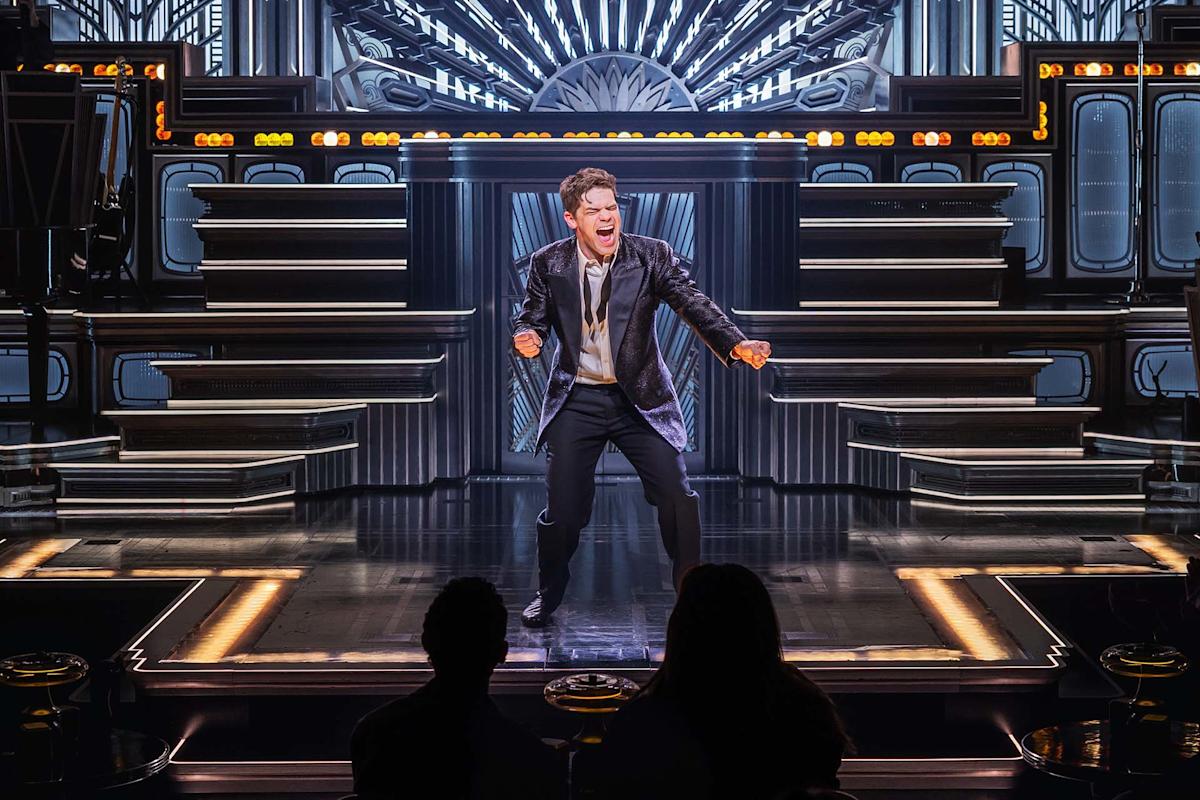 A performer in a black jacket sings energetically on a futuristic, illuminated stage, with two audience members silhouetted in the foreground.
