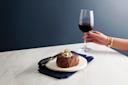 A person holds a glass of red wine next to a thick steak topped with a creamy sauce, placed on a white plate with a dark blue napkin, on a marble table against a dark gray background.