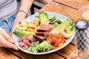 A person holds a plate of salad with seared tuna slices, lettuce, cherry tomatoes, hard-boiled egg, roasted potatoes, cucumbers, pickled onions, and green beans on a wooden table.