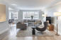 A modern living room with a gray sofa, a dark armchair, and a round coffee table on a beige rug. Large windows let in natural light, and there's a dining area with chairs in the background. A floor lamp stands beside the sofa.