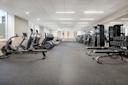 A spacious, modern gym with cardio machines like treadmills and ellipticals on the left, weightlifting equipment and racks of dumbbells on the right, and large windows letting in natural light.