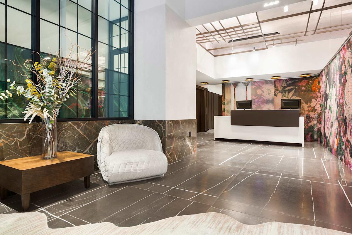 A modern hotel lobby with marble floors, a white armchair, a wooden table with a decorative vase, large windows, and a reception desk in the background. The walls have floral and marble patterns.