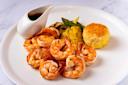 A plate of grilled shrimp with a side of roasted vegetables, including corn and asparagus, and a round portion of mashed potatoes with a crispy top. There is also a small gravy boat with dark sauce.