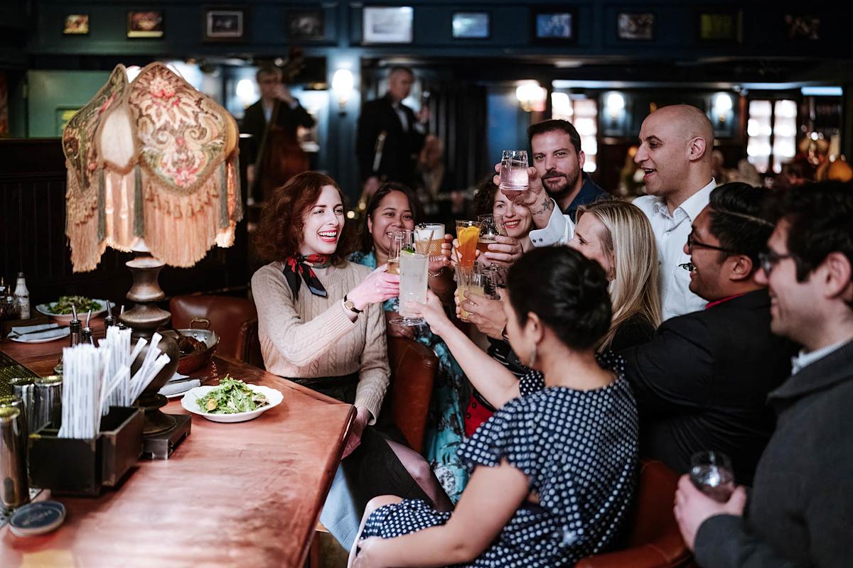A group of people sit around a table in a cozy bar, raising their glasses in a cheerful toast. They are smiling and appear to be enjoying each other's company. A vintage lamp and plates of food are on the table.