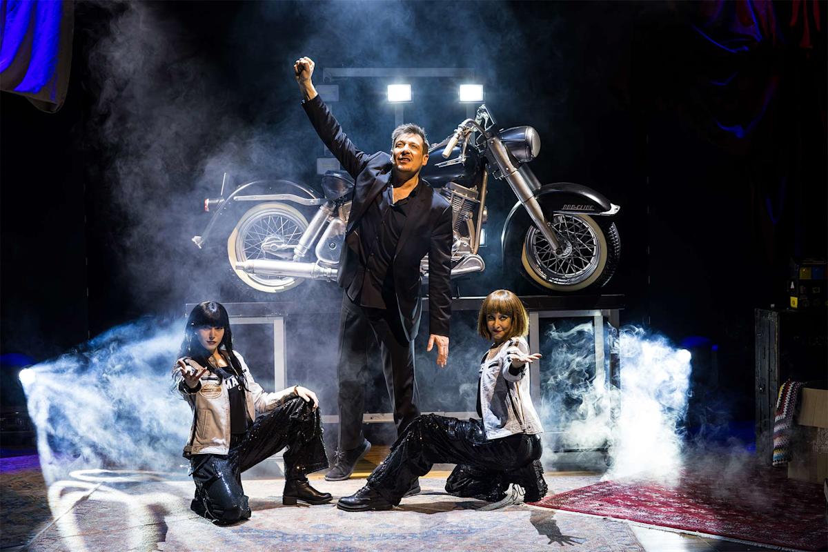 A man in black stands confidently with one arm raised between two crouching women on a smoky stage, with a large motorcycle displayed behind them.