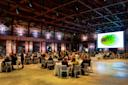 A large event hall with people seated at round tables. Graffiti decorates the walls, and a big screen displays a watermelon image. The atmosphere is lively, with attendees engaged in conversation under a high ceiling with exposed beams.
