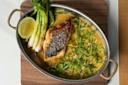 An oval dish with a seared fish fillet on creamy sauce, topped with sliced green chili and herbs. Garnished with grilled green onions and a lime wedge, set on a wooden board.