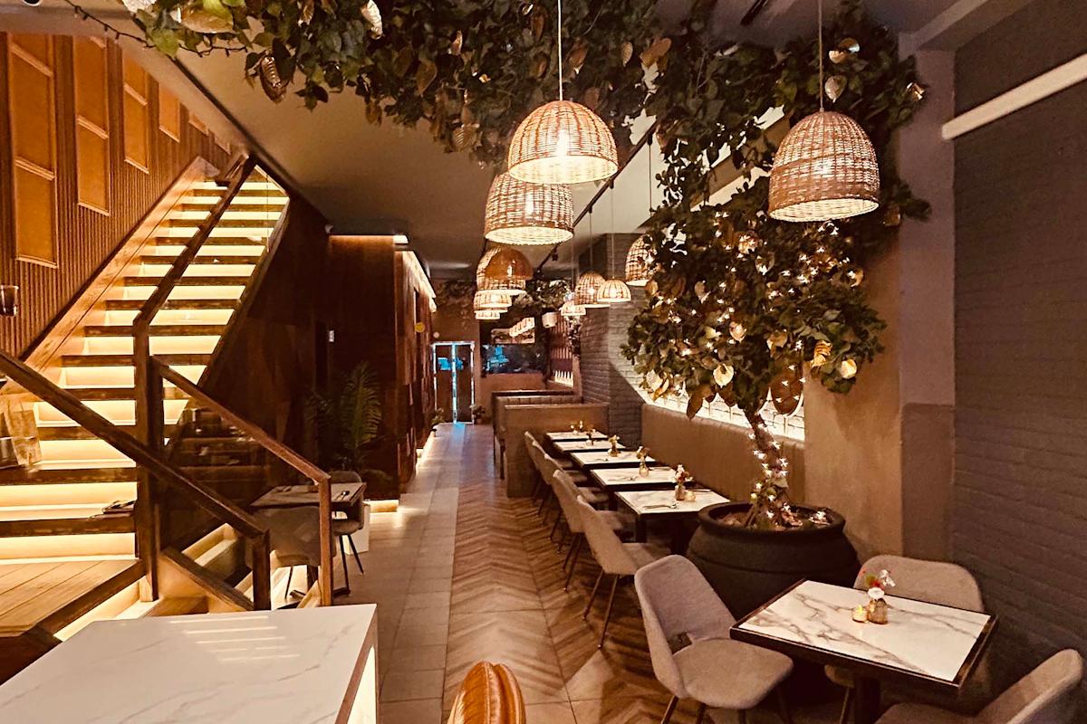 A cozy restaurant with hanging wicker lights, potted trees with string lights, marble tables, cushioned chairs, and a wooden staircase on the left. The atmosphere is warm and inviting.