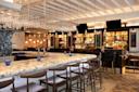 A stylish bar with a marble countertop and wooden barstools. Shelves stocked with bottles are visible in the background. Hanging wine glasses and pendant lights add a touch of elegance. The floor features a geometric pattern.