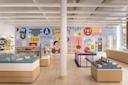 A bright, modern store with wooden floors, displays of books and gifts, and a colorful mural featuring abstract faces, letters, and New York-themed illustrations on the back wall.