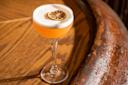 A coupe glass with an orange cocktail topped with a layer of foam and garnished with a dried citrus wheel, placed on a wooden surface.