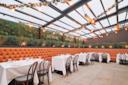 Stylish restaurant interior with a glass roof, featuring rows of tables with white tablecloths and brown chairs. The walls are adorned with greenery, and warm string lights hang above, creating a cozy ambiance.
