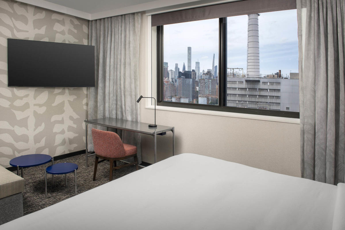 Hotel room with a bed, a wall-mounted TV, a desk, and a chair. Large window offers a city view with skyscrapers and industrial elements visible. Neutral tones with a patterned wall add a modern touch.