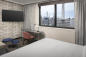 Hotel room with a bed, a wall-mounted TV, a desk, and a chair. Large window offers a city view with skyscrapers and industrial elements visible. Neutral tones with a patterned wall add a modern touch.