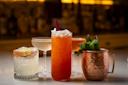 A lineup of four assorted cocktails in different glasses sits on a bar, including a salted-rim drink with lime, a coupe glass with a pale cocktail, a tall orange drink with ice and garnish, and a copper mug with mint leaves.